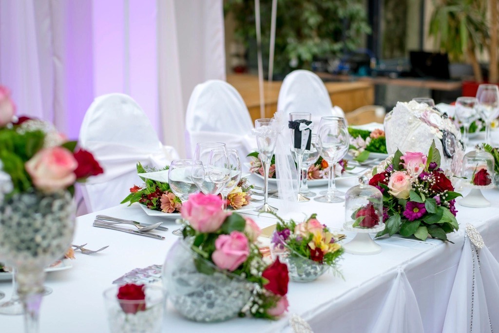 Mesa de festa com arranjos de rosas, taças e cadeiras com capa branca