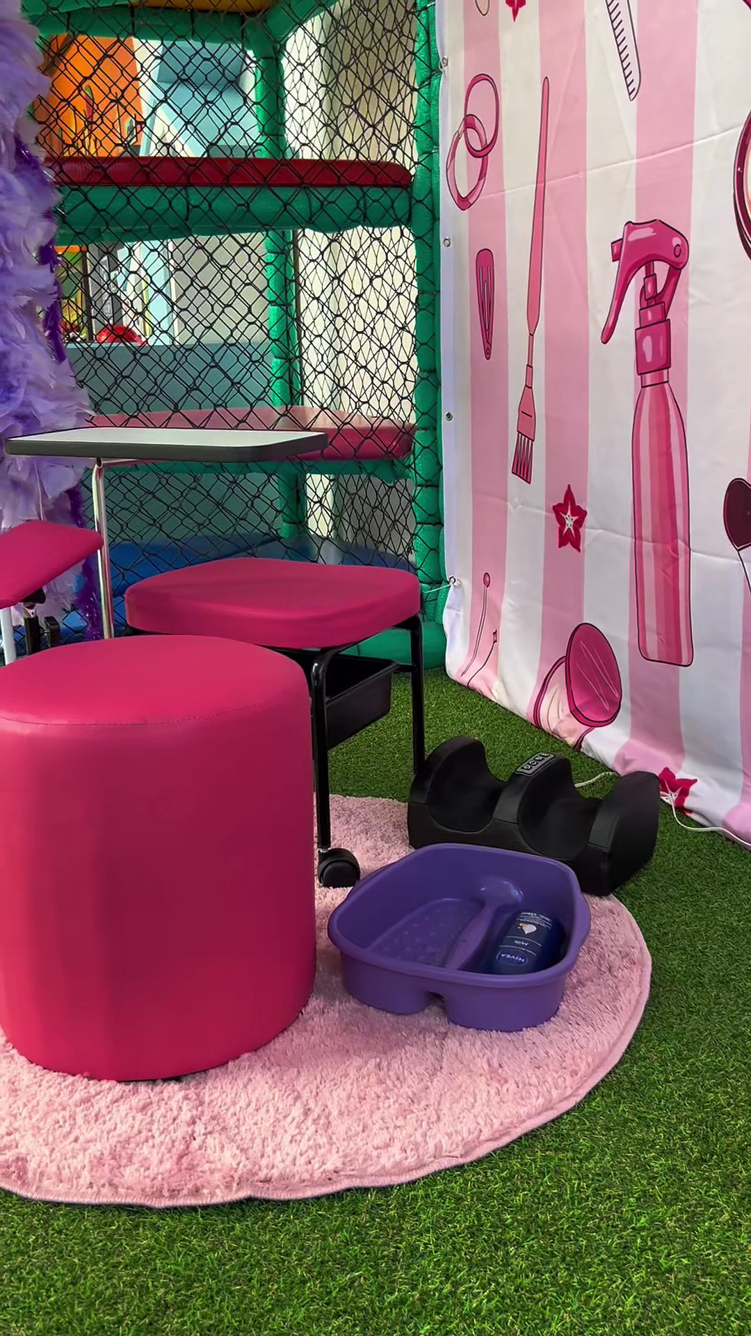 Cantinho de beleza da casa de festas: pufes rosa, carrinho de manicure, spa para os pés e painel decorado com desenhos de secador e escova, sobre grama sintética.