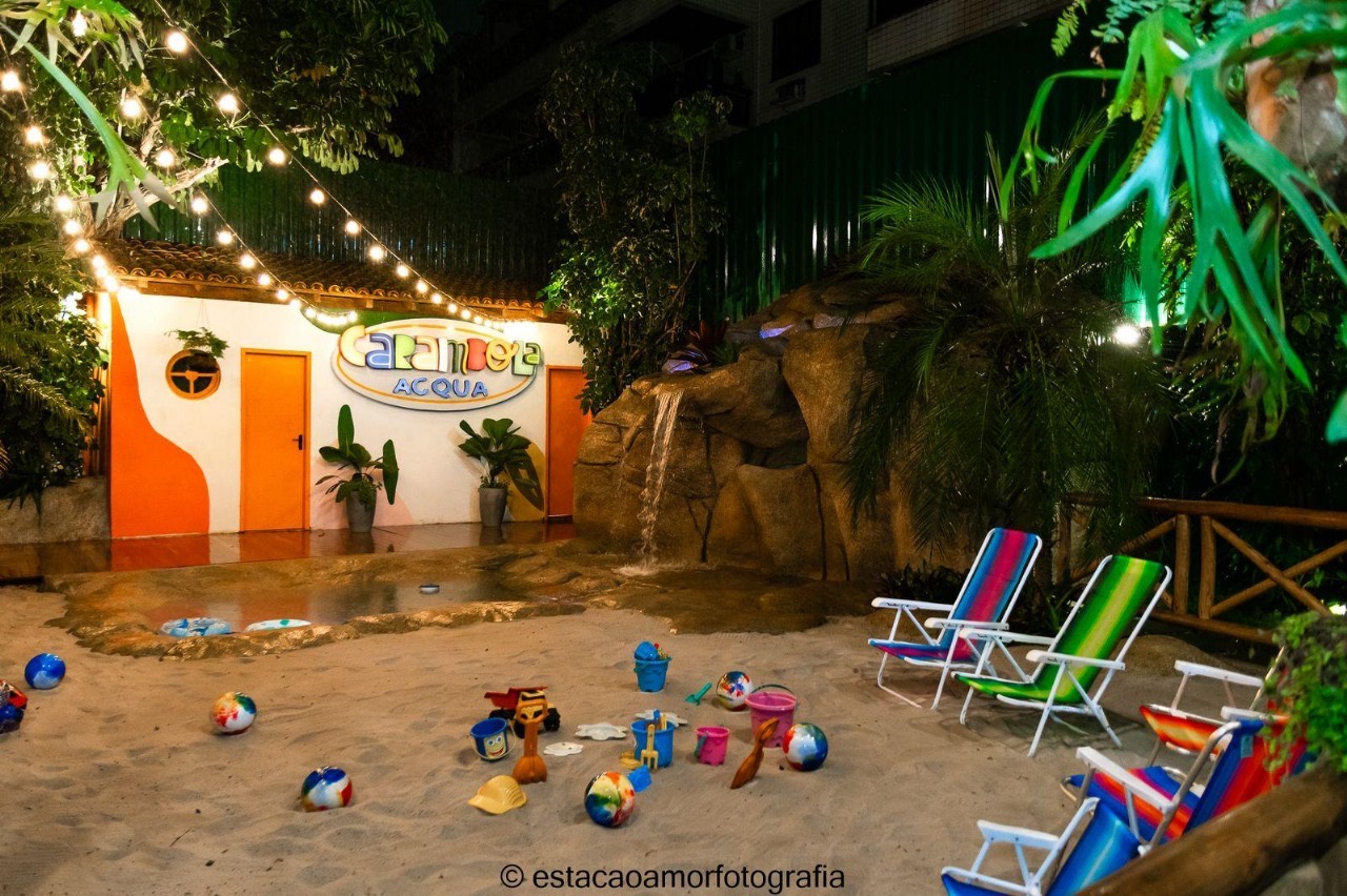 A prainha à noite, iluminada por varal de luzes: areia com baldinhos e bolas, espreguiçadeiras listradas e a cascata iluminada sobre as pedras.
