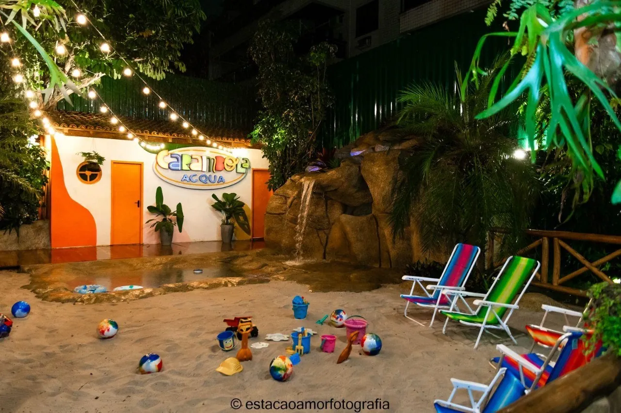 A prainha à noite, iluminada por varal de luzes: areia com baldinhos e bolas, espreguiçadeiras listradas e a cascata iluminada sobre as pedras.